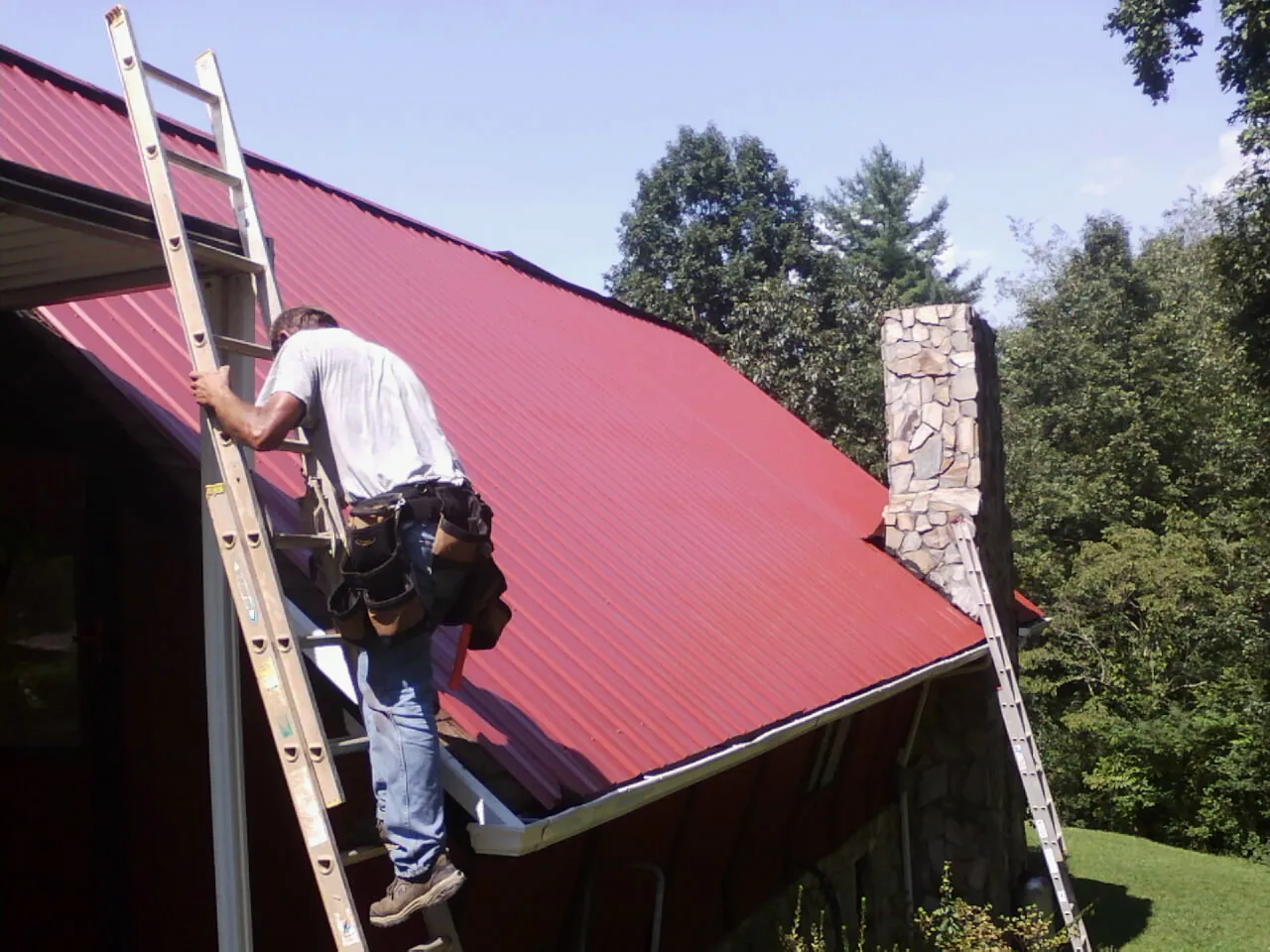 Roofer on a ladder beside a red metal roof and stone chimney — Soffit & Fascia Repair by Roofing Brothers in Ottine